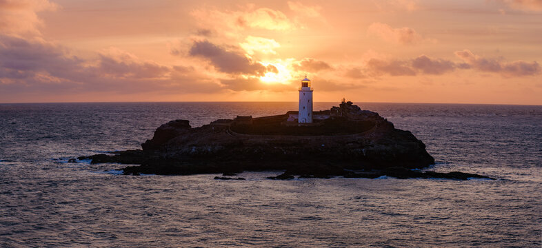 View of Godrevy Lighthouse on a rocky island during a fiery sunset with orange clouds over the Atlantic Ocean in Hayle, Cornwall, United Kingdom.