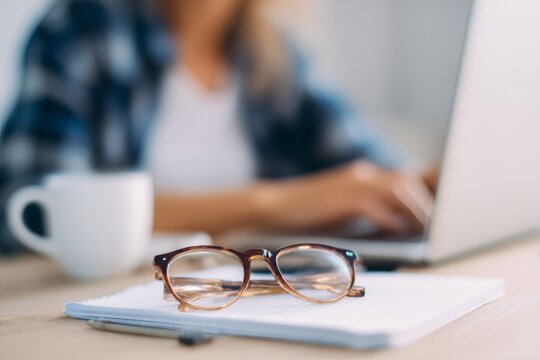 Close up of eyeglasses on notepad with blurred woman working on laptop. Freelancer typing at home office desk. Remote work and vision concept
