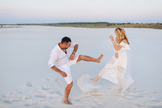 A young man swings his leg and plays with the sand on the beach next to a beautiful blonde woman. White shirt, dress and shorts. The concept of love, vacation and honeymoon.