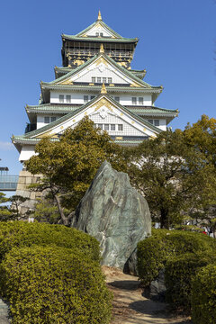 View of the historic Osaka Castle main tower with its white walls and green roofs rising above lush trees and a large decorative rock under a clear blue sky in Osaka, Osaka, Japan.