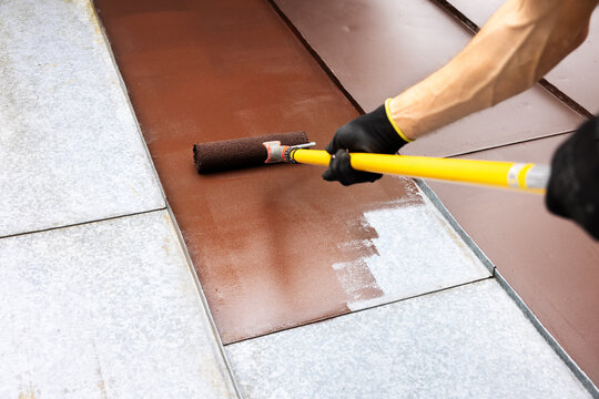 worker painting metal roof with roller and brown paint. house roofing restoration