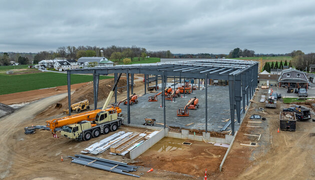 Workers are building a new structure in a rural area. Heavy machinery is nearby, and the site shows steel beams and equipment. The sky is overcast as work continues on the project.