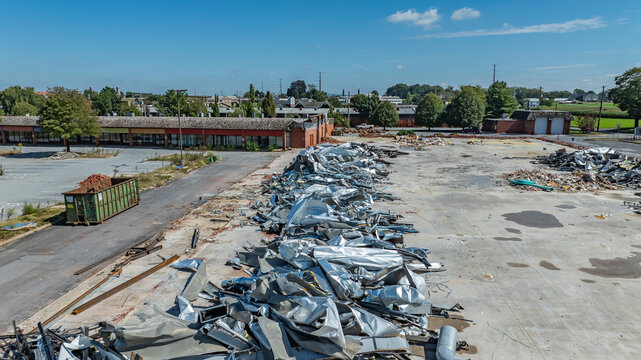 The scene shows a large area of a former factory site with metal debris scattered across the ground. The sky is clear with a few clouds. Green fields and buildings are visible in the background.