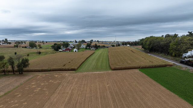 Cornfields line the land with a few houses visible in the distance. A railway track runs along the edge of the fields as clouds fill the sky. The scene shows rural life on a fall day.