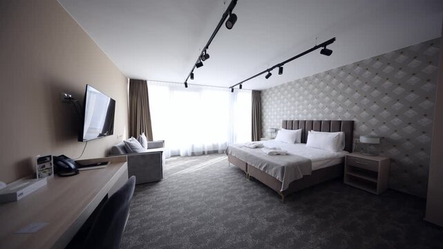 Interior shows twin beds, upholstered headboard, accent wall, TV, desk, sofa, sheer curtains. Bright daylight, slow lateral pan, wide angle, black track lights.