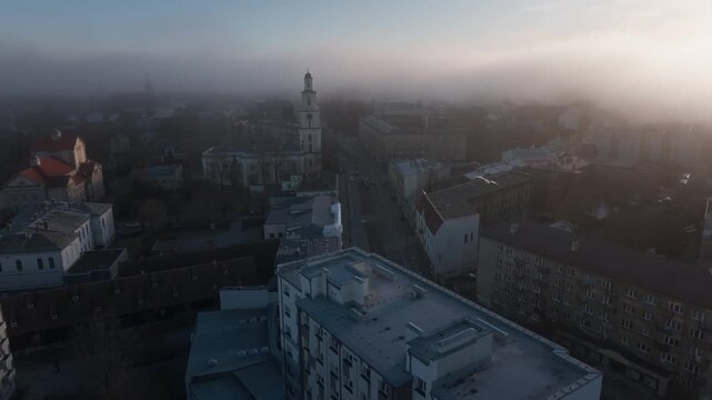 Aerial view shows central district with multi tiered church bell tower and long straight avenue. Soft morning fog drifts as sun glare right adds warm backlight.
