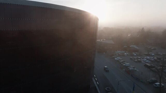 Aerial drone pans past a large brick clad arena with metal ribbed roof by a busy parking lot at sunrise in a town center, with a car moving and warm lens flare.