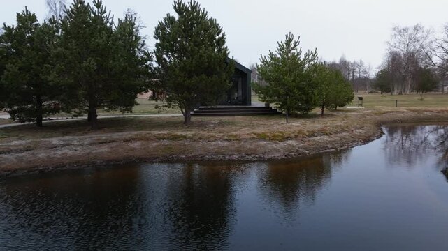 Small black cabin with glass front on peninsula beside reflective pond. Aerial lateral drone motion reveals calm ripples, grill, pathway, pines, birch, winter light.