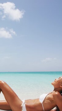Woman relaxing on summer beach vacation. Sexy young Asian Caucasian adult in bikini sitting down on perfect white sand with sky copy space for tropical travel destination concepts. Vertical video