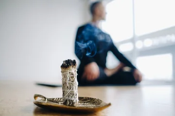 Fototapeten Lotusblume A male athlete in black sportswear sits in the lotus position against the background of an incense candle in the gym.  © Alex