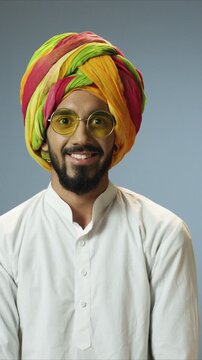 Cheerful close up portrait of Indian handsome happy young man wearing colorful turban and round glasses smiling at camera. Male standing relaxed and showing joy. Indoor lifestyle concept.