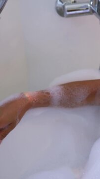 Vertical video: Settling bare forearm on tub rim in bathroom, relaxing in bubble foam with faucet