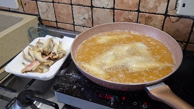 Small fresh fish deep frying in hot boiling oil inside frying pan preparing traditional delicious mediterranean seafood meal on stove in kitchen in Italy.
