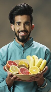 Smiling close up portrait man holding bowl of fresh fruit and presenting healthy snack. Male offering bananas citrus and kiwi. At home. Indoor lifestyle food concept nutrition focus balanced diet idea