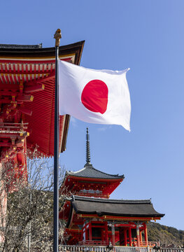 View of the Japanese flag waving in front of the historic Kiyomizu-dera temple with its iconic red architecture and a clear blue sky in Kyoto, Kyoto, Japan.