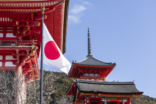View of the Japanese flag waving in front of the red wooden architecture of Kiyomizu-dera temple with a pagoda spire under a clear blue sky in Kyoto, Kyoto, Japan.