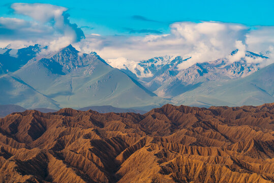 View of Ak Say Canyon with its unique eroded brown ridges and snow-capped peaks under a bright blue sky with scattered clouds in Ak Say, Issyk-Kul Region, Kyrgyzstan.