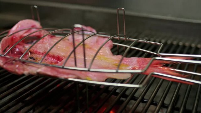 Fresh red snapper grilling inside a metal basket on a hot barbecue. Close up of the raw seafood cooking over glowing charcoal in a restaurant