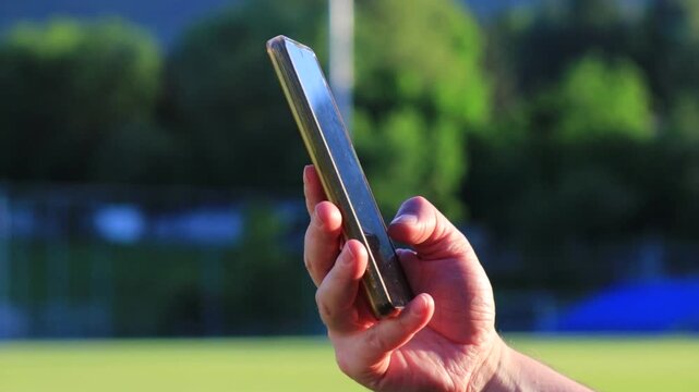 Male hand scrolling content on smartphone near football stadium field at sunset, close up of man using mobile phone browser outdoors, social media browsing, mobile internet, online communication