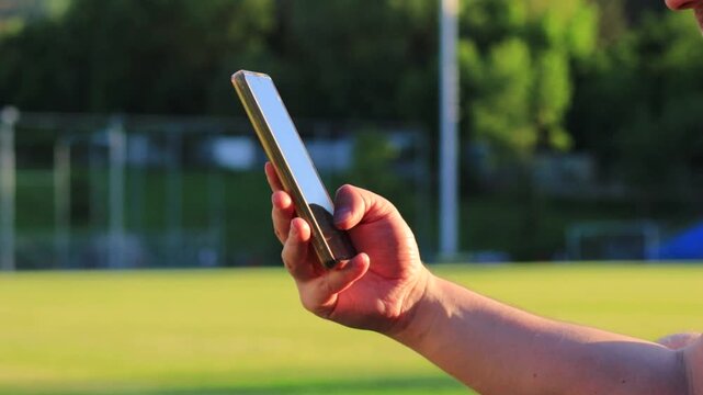 Male hand scrolling content on smartphone near football stadium field at sunset, close up of man using mobile phone browser outdoors, social media browsing, mobile internet, online communication