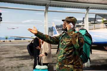 Naklejka premium Airport traveler wearing military clothing talking on smartphone, doing hand gestures, army soldier in modern airport terminal carrying backpack, doing phone call, for travel planning.