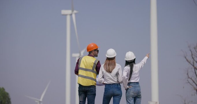 A team of three engineers in hardhats and safety vests review blueprints and discuss a renewable energy project while inspecting wind turbines and planning wind farm development outdoors.
