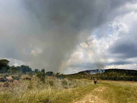 View of a large plume of smoke rising from a wildfire on a hillside with a firefighter walking along a dirt path Cape Town, Western Cape, South Africa.
