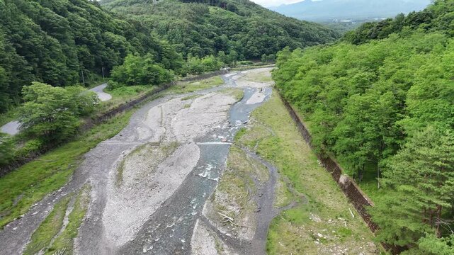 Aerial view reveals a winding riverbed through forested hills. Shallow water flows through rocky, dry patches. Green hills flank the river's path on both sides