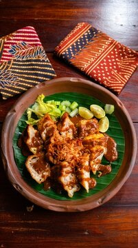 Fried fish dumpling batagor served with thick peanut sauce, crispy shallots, lime wedges and celery on banana leaf in a clay bowl, with batik fabric background.
