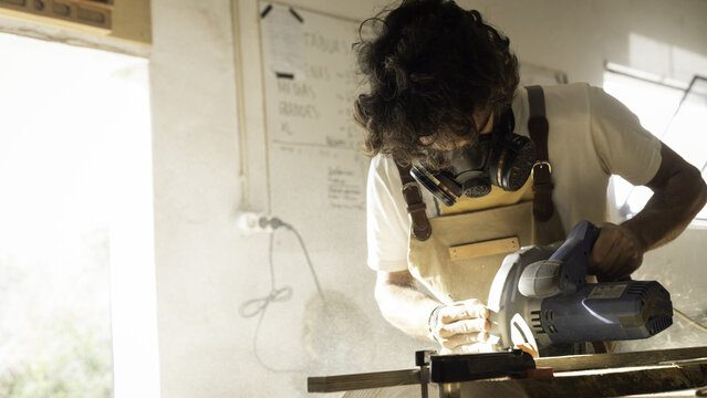 View of a woodworker wearing a protective mask using a circular saw to cut a wooden plank in a workshop in Lisbon, Lisbon, Portugal.