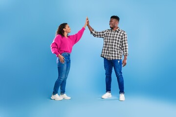 Fototapeta premium Joyful Emotion. Happy Young Black Man And Woman Giving High Five While Standing In Studio With Light Blue Background. Celebrating Success Together In A Fun And Energetic Atmosphere.