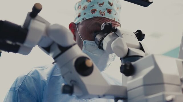 Surgeon in blue scrubs and patterned cap focuses on surgical microscope during eye procedure, with medical equipment and bright operating room background visible