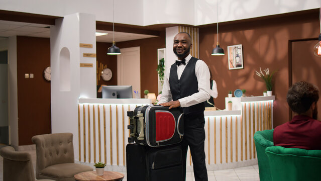 African American bellboy takes suitcases to help important guest with room accommodation, providing luxurious concierge services in hotel lobby, employee wecoming and assisting businessmen