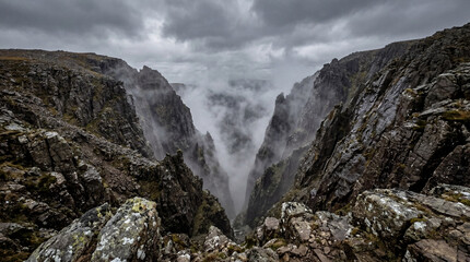 Fototapeta premium Looking Down Into Steep Rocky Ravine With Swirling Mist Between Sharp Stone Walls Mountain Landscape