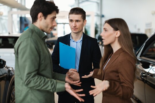Focused Expressions On Faces Of Young Adults. Two Men Caucasian And One Woman Negotiating And Discussing Car Options In Modern Dealership Office Setting. Business Concept.