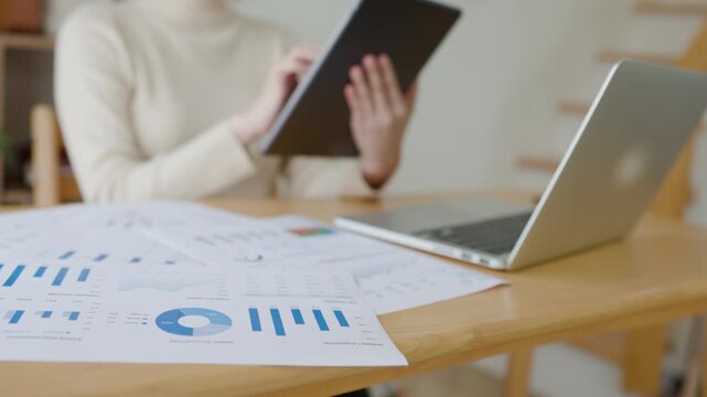 Printed charts on a desk beside a laptop, while a person reviews data on a tablet, suggesting business analysis and reporting