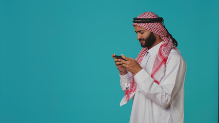 Happy person having fun with game on his smartphone device, wearing traditional arabic attire, in the studio, muslim model enjoying e sport tournament with mobile phone videogames. © DC Studio