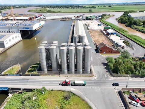 Aerial view of industrial metal silos next to a canal with a tractor pulling a trailer on the road, surrounded by green fields and waterways in Goole, England, United Kingdom.