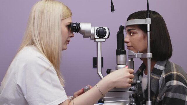 Female optometrist examines a woman's eye using a slit lamp in a clinical setting with purple walls and medical equipment visible in the background