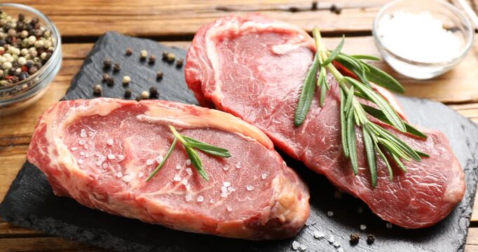 Raw beef steaks with salt, rosemary and spices on wooden table, closeup. Camera moving right