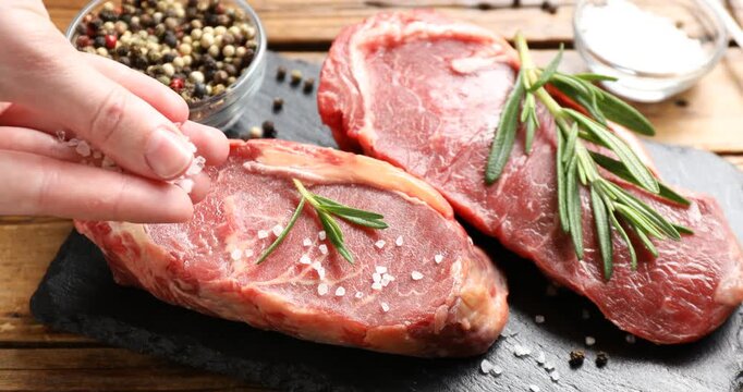 Woman adding salt to raw beef steak at wooden table, closeup