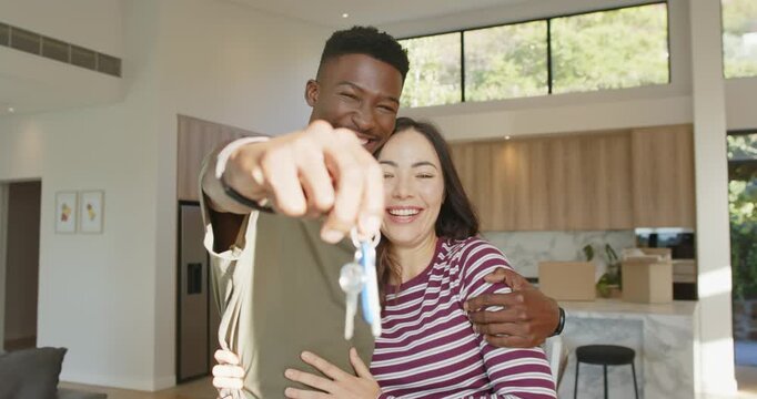 Diverse couple embracing and presenting keyring after receiving keys at open-plan home