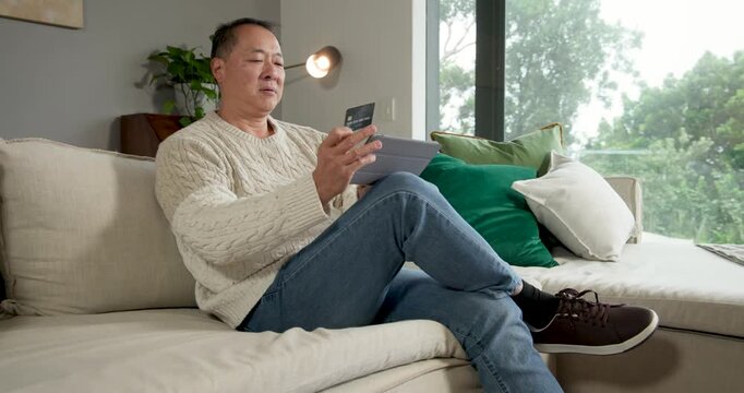 Senior Asian male sitting on sofa at home, holding tablet and card, reading card, entering payment