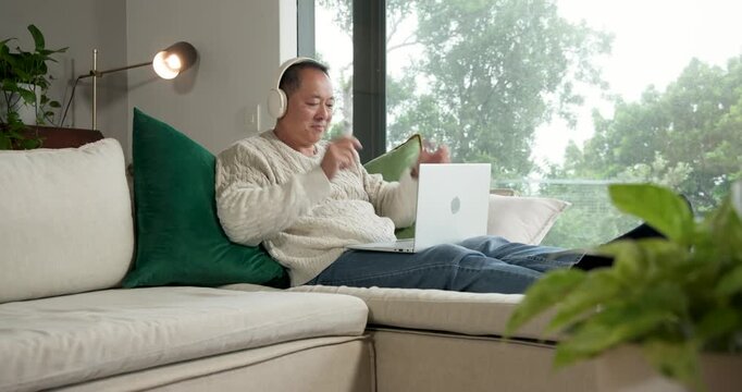 Senior Asian man lifting headset, donning headphones, typing on white laptop at home working