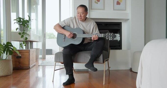 Senior Asian man sitting on chair at home, strumming grey acoustic guitar, practicing and smiling