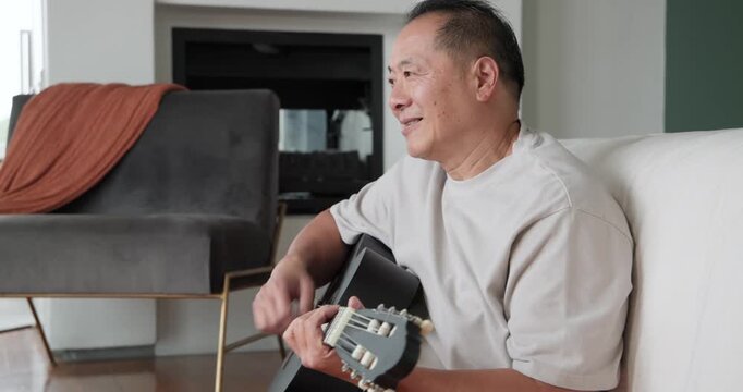 Senior Chinese man lifting black acoustic guitar onto lap practicing chords on couch, copy space