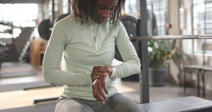African woman checking smartwatch, adjusting strap, tracking workout at gym bench wearing mint top