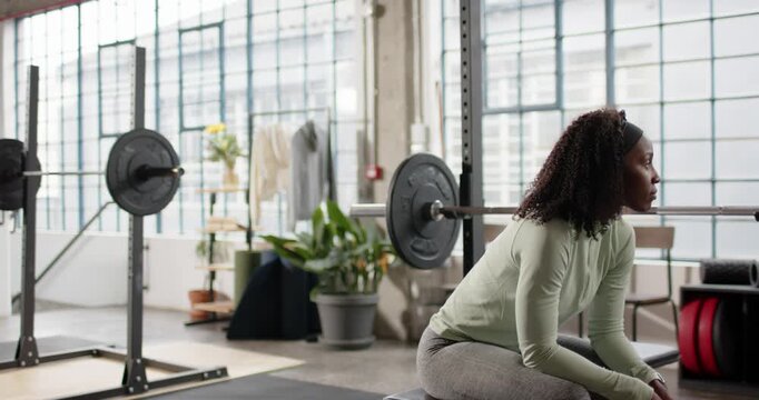 African American woman sitting on bench in gym in green leggings preparing set with barbell