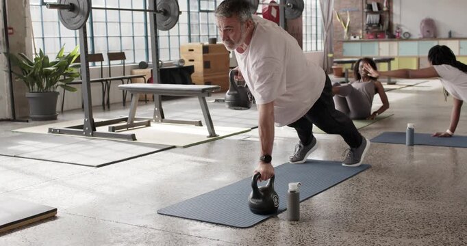 Diverse group on class cue in loft studio as middle-aged man doing kettlebell-plank rows for core
