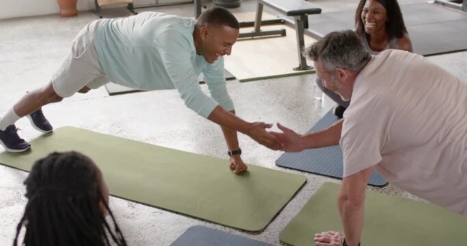 Diverse fitness class doing plank African American man blue top tapping partner on mats for balance